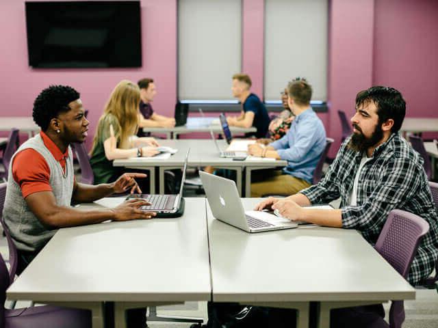 SBU学生坐在教室里的桌子旁，拿着笔记本电脑